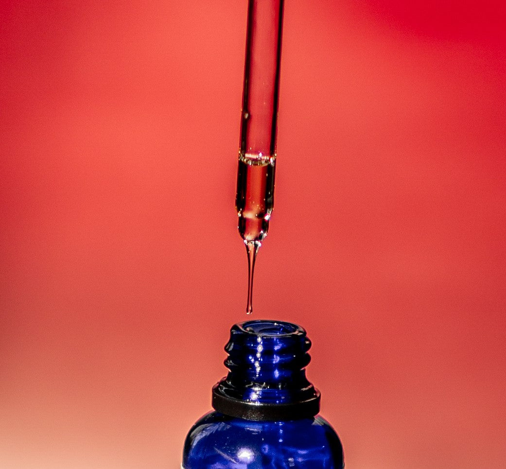 A dropper filled with prickly pear seed cactus oil dropping the oil into a blue glass bottle amongst a red background