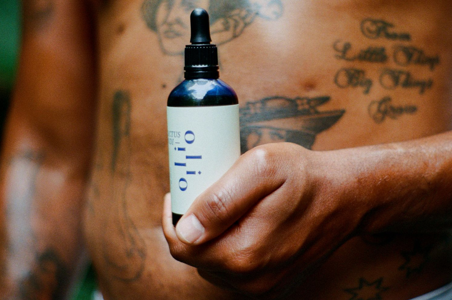 Tattooed person holding a bottle of body oil labeled 'oli oli' with tattoos on their arm and body.