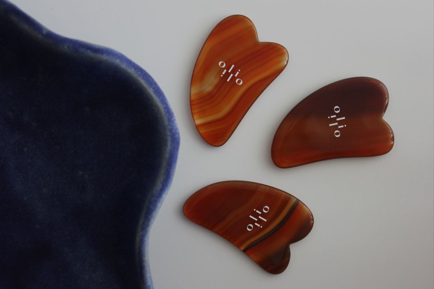 Three warm onyx stone gua sha tools on a white surface with a blue ceramic bowl