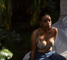 dark skin female model sitting outside amongst greenery in her bra and blue jeans with her face looking at the camera while she has on prickly pear cactus face oil on her face and neck and prickly pear cactus body oil on her chest stomach and arms